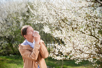 woman with seasonal allergies blowing her nose into a tissue next to a tree with white buds
