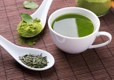 cup of green tea next to spoonfuls of dried green tea leaves and green tea powder