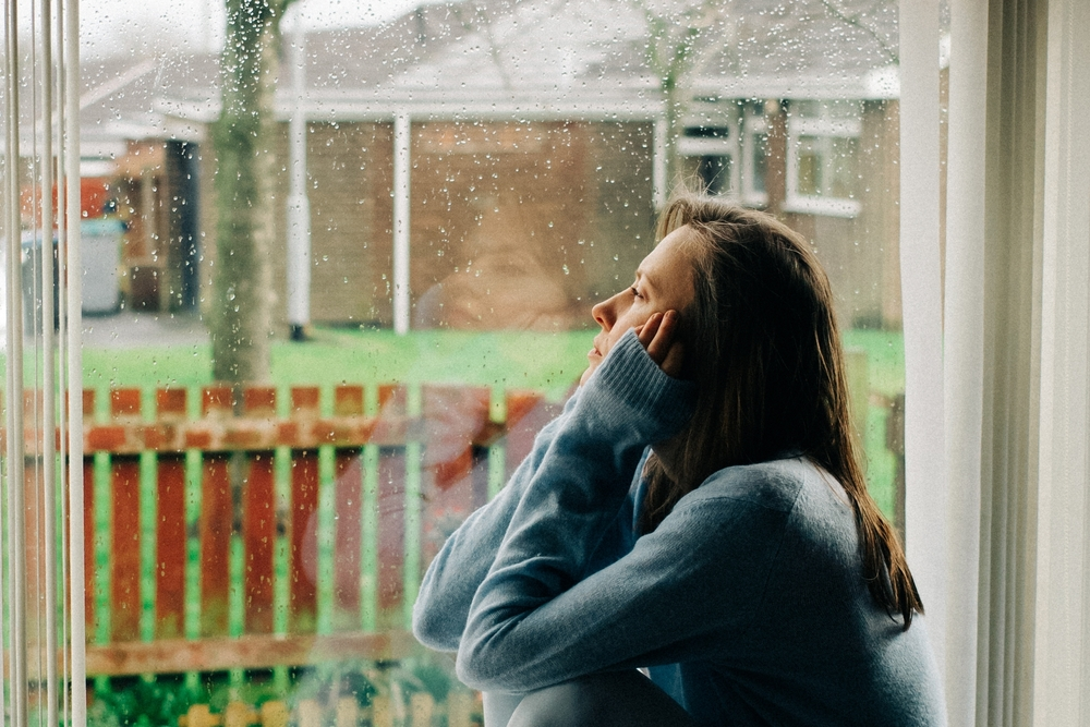 depressed woman looking out window at cold, rainy day